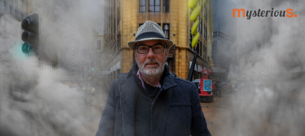 Nick walks through the fog. Nick is walking through the fog. He is wearing warm clothes and has a beard, glasses and a hat.